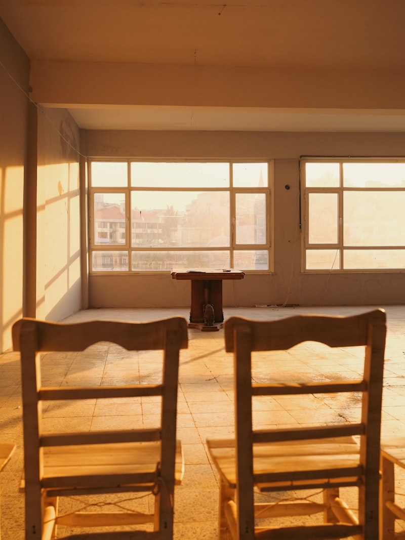 Dos sillas de madera frente a una mesa, en una sala iluminada por ventanales.