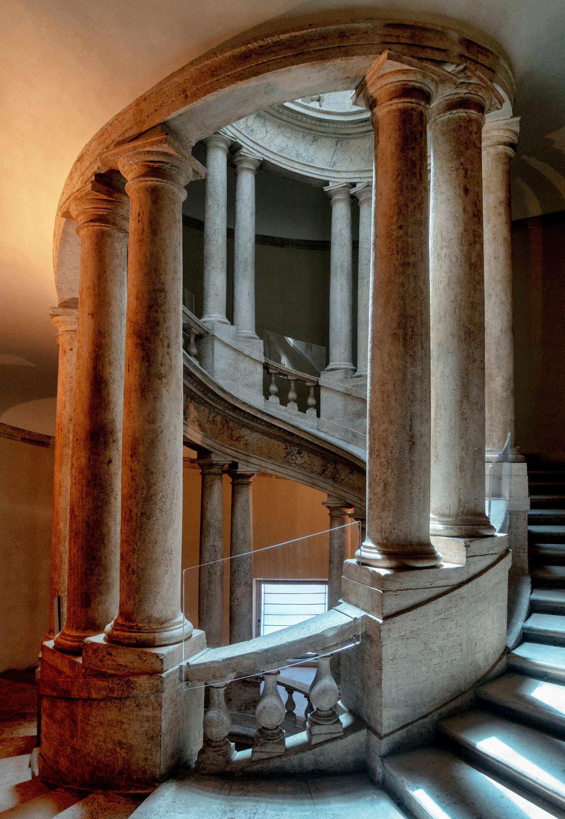Escalinata de mármol en espiral enmarcada por columnas clásicas, iluminada por una claraboya cenital.